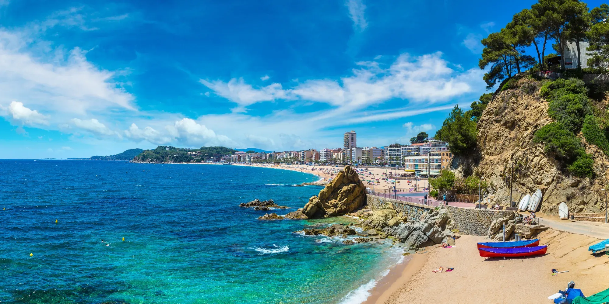 Playa costera con barcas, acantilados rocosos y edificios de pueblo junto al mar.