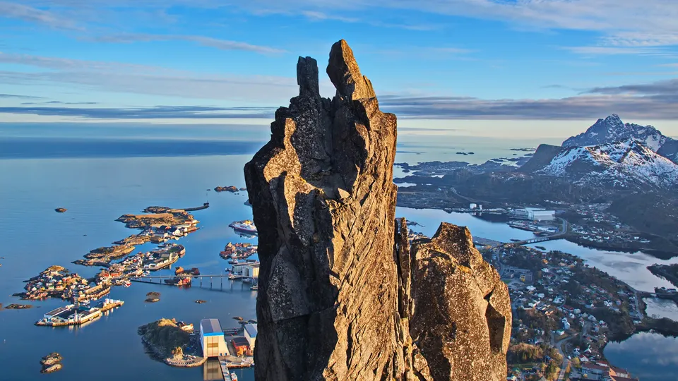 Dramatisk fjellspir som reiser seg over kystby med havn og snøkledde fjell.