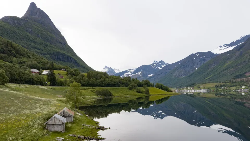 Fjellsjø som reflekterer topper og skogskledd dal med jordbruksland og båthus.