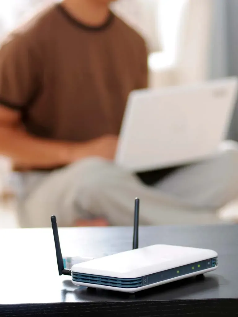 Router WiFi en primer plano con persona usando portátil de fondo.