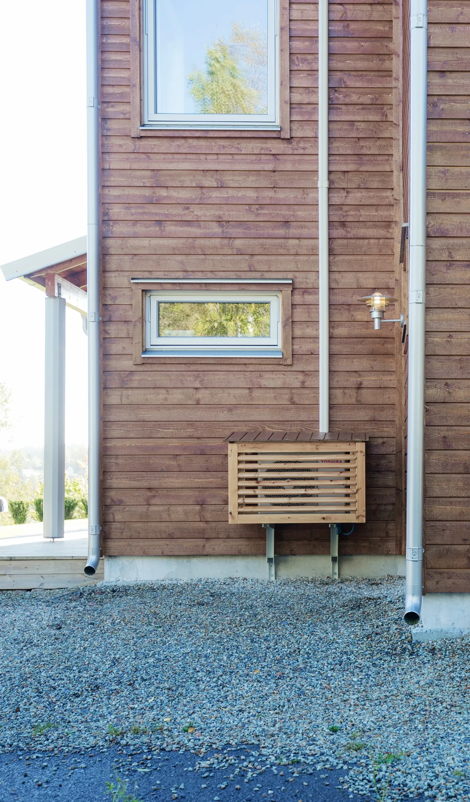 Outdoor unit of a Toshiba heat pump mounted on the exterior wall of a wooden house, enclosed in a decorative wooden casing on a gravel surface.