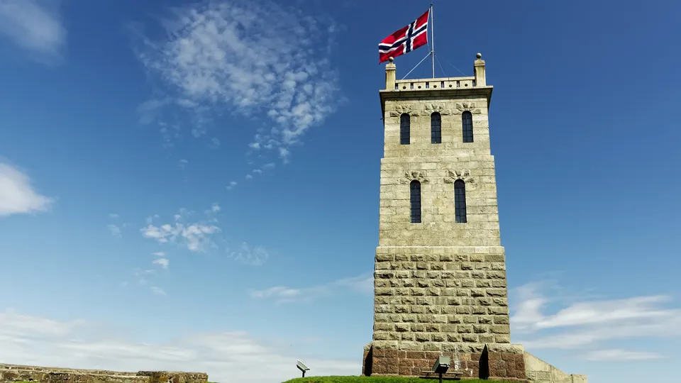 Steintårn med norsk flagg som vaier mot blå himmel.