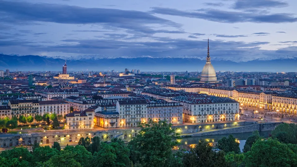 Paesaggio urbano di Torino al tramonto con la Mole Antonelliana illuminata e le Alpi innevate sullo sfondo.