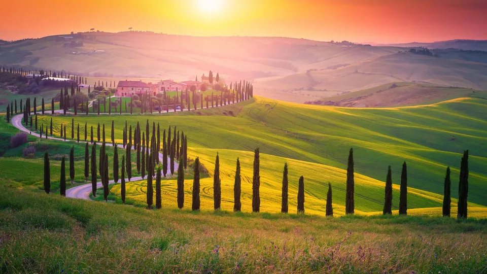 Villa toscana con viale di cipressi al tramonto su colline ondulate.