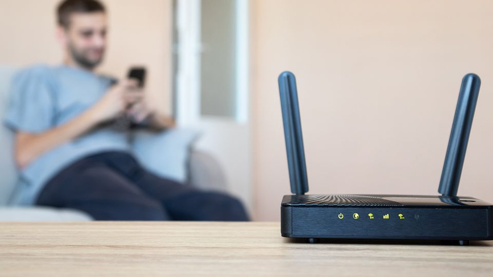 Bilde av en router som står i forgrunnen mens en person sitter på mobilen i en sofa i bakgrunnen.