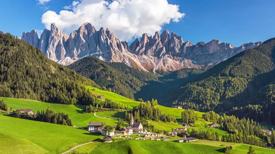 Villaggio alpino con chiesa in una valle verde circondata da prati e montagne boscose.
