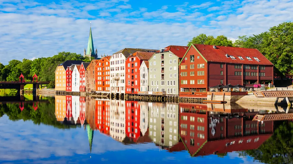 Fargerike trehus reflektert i stille elvevann med katedral-spir i bakgrunnen.