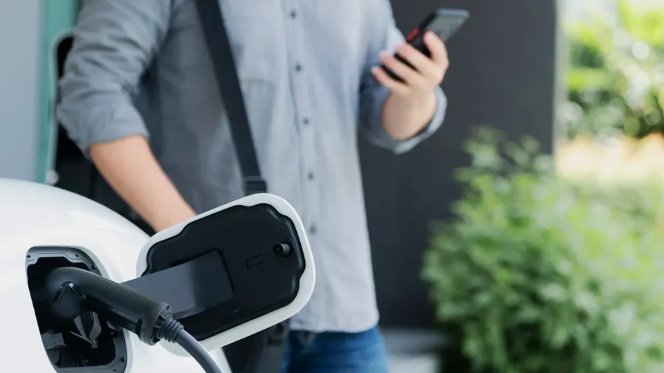A person checking their phone in the background while an EV charges in the foreground.