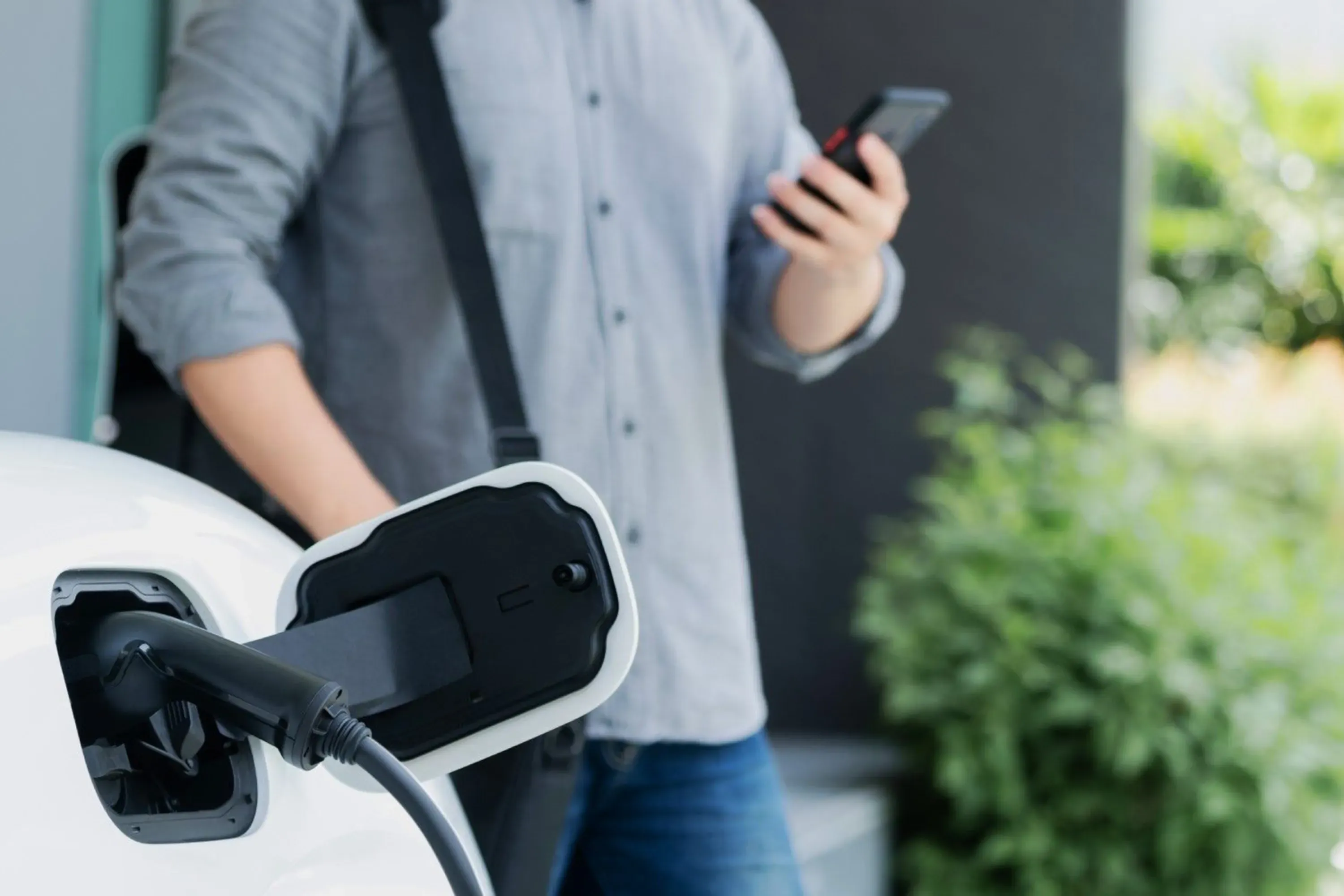A person checking their phone in the background while an EV charges in the foreground.