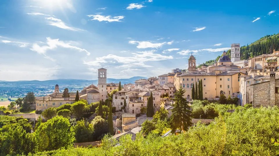 Borgo medievale su collina con edifici in pietra e torri di chiese che si affacciano sulla valle verde.
