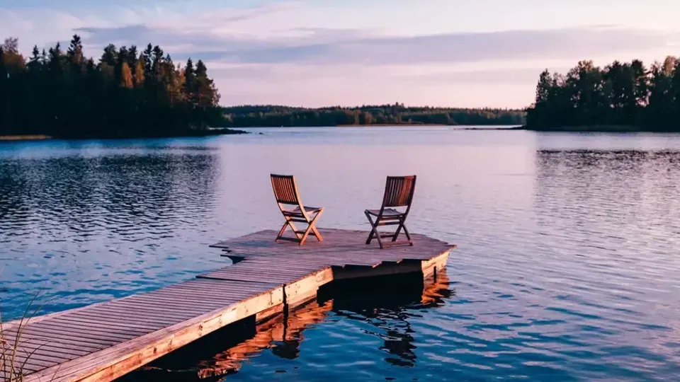 Två stolar på ett trädäck som sträcker sig ut i en lugn sjö omgiven av skogbevuxen strand.