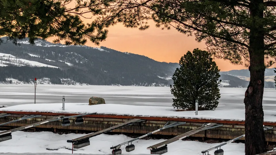 Frossen innsjø med snødekt båtbrygge og furutrær ved solnedgang.