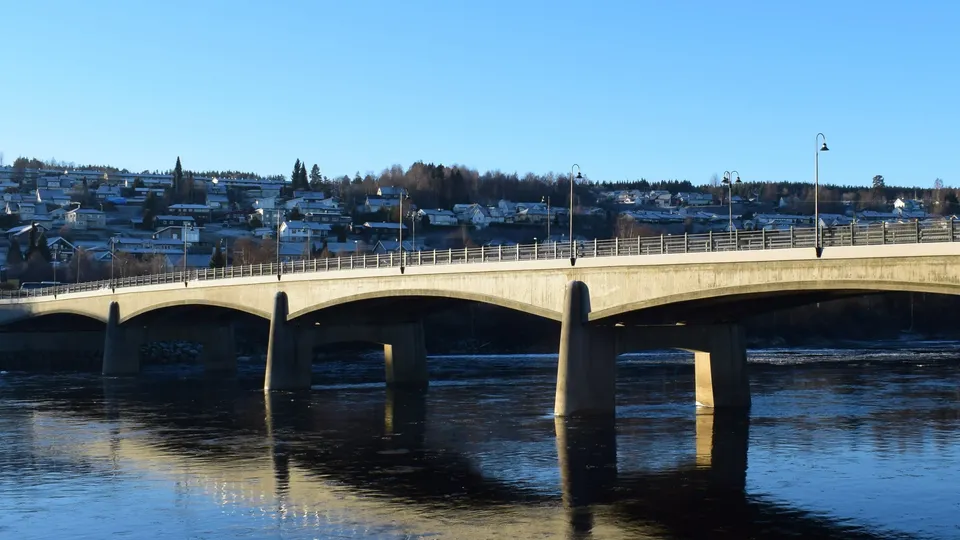 Betongbru med metallrekkverk som spenner over en elv, bolighusen synlige på bakken bak.