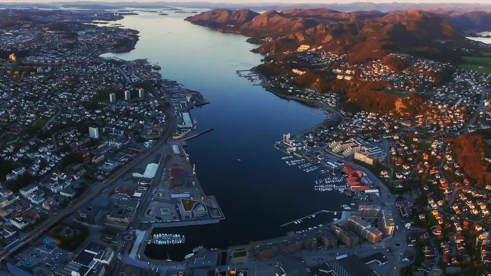 Luftfoto av kystbyen Sandnes med havn og fjell over blått vann ved solnedgang.