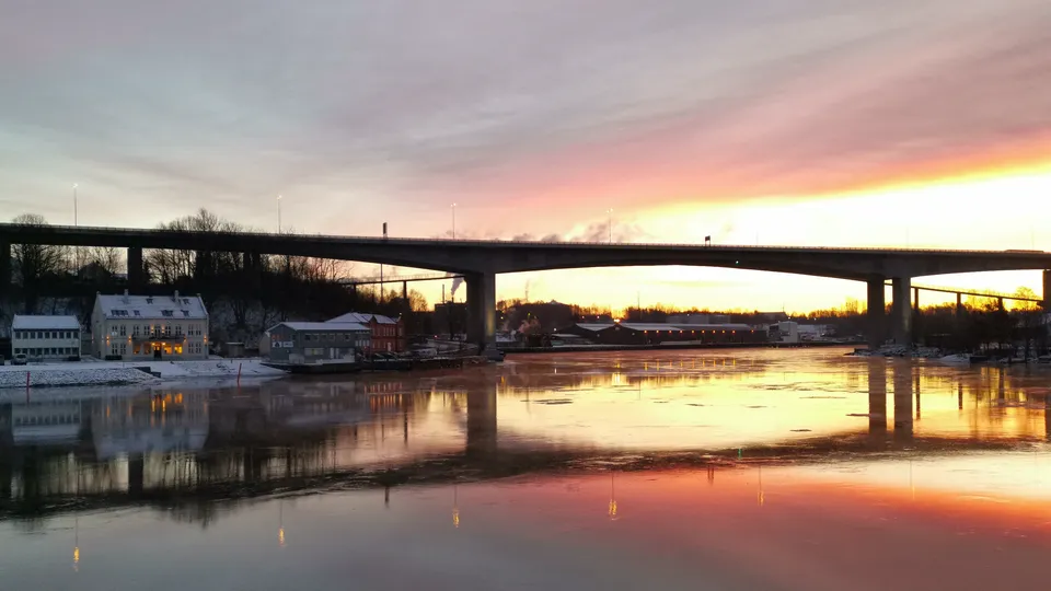 Solnedgang over elv med bro og bygninger ved vannkanten reflektert i vannet.
