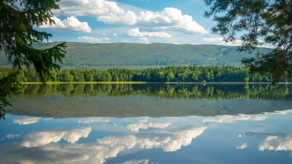 Stille innsjø i Steinkjer som reflekterer skogkledd fjell og skyet himmel.