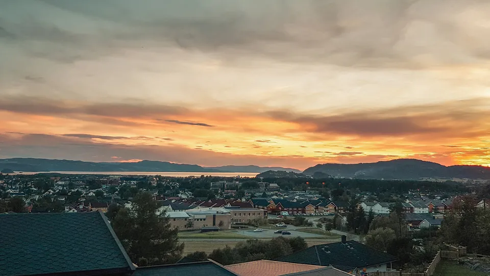 Forstadsbebyggelse ved solnedgang med fjell og vann i bakgrunnen.