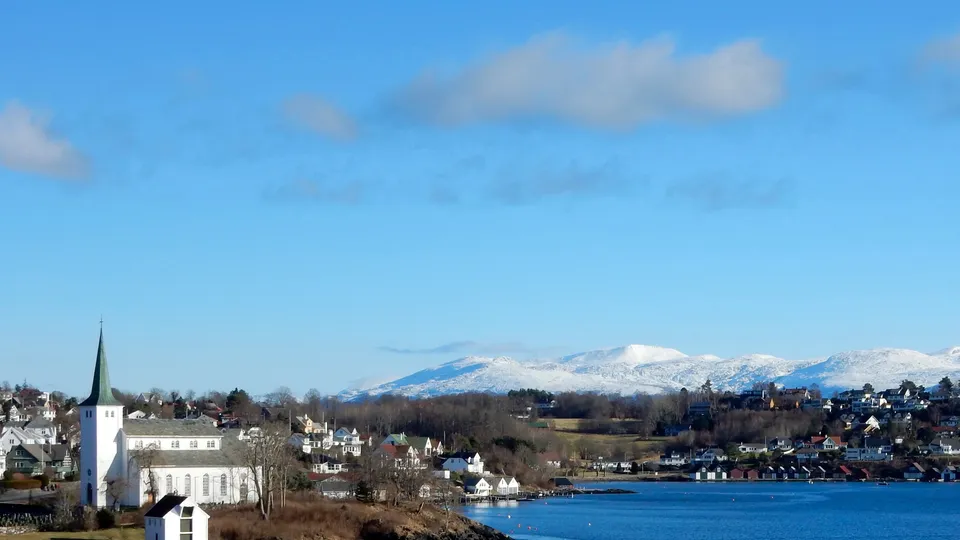 Hvit kirke på steinete halvøy på Stord med utsikt over blå fjord og snødekte fjell i bakgrunnen.
