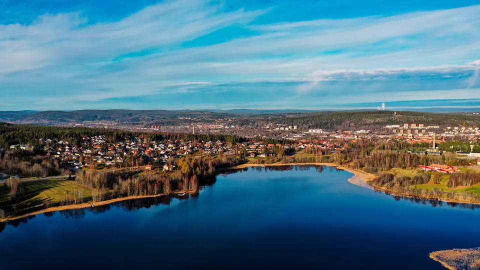 Flygfoto över sjö omgiven av skog och bostadsområden med en stadsskyline i fjärran.