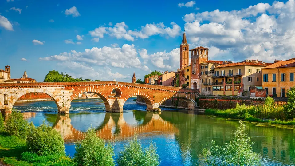 Antico ponte in mattoni che attraversa il fiume con torre della chiesa e edifici colorati sullo sfondo.
