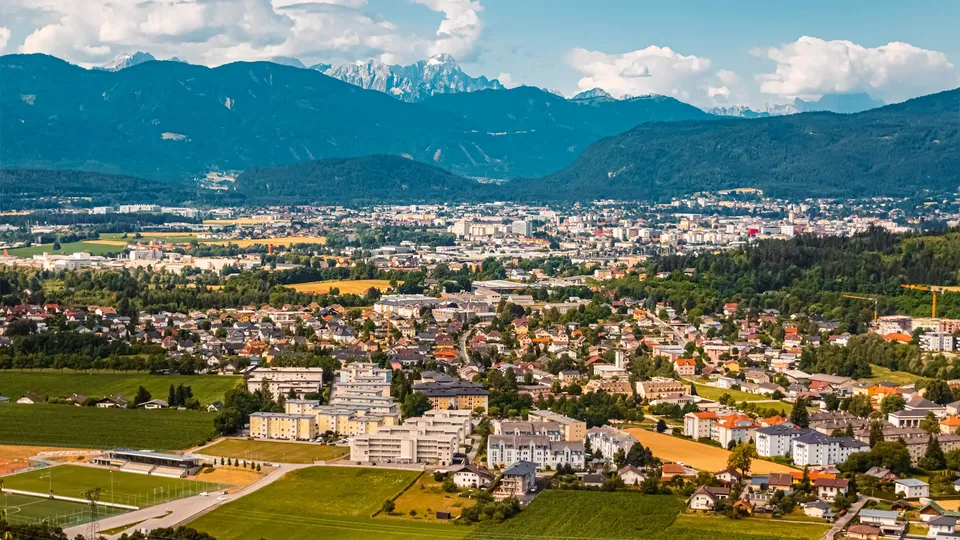 Luftaufnahme von Villach in Kärnten im Talkessel, umgeben von Wohnhäusern, Feldern und den Alpen im Hintergrund unter blauem Himmel mit Wolken.