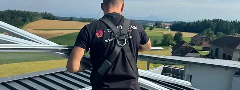 OH Voltaik Solaranlagenbau GmbH – Monteur mit Sicherheitsgeschirr auf Blechdach mit Blick auf ländliche Landschaft