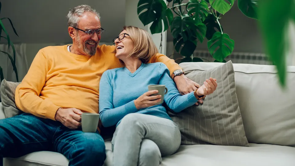 Ældre mand og kvinde sidder på sofa, holder kopper og smiler til hinanden.