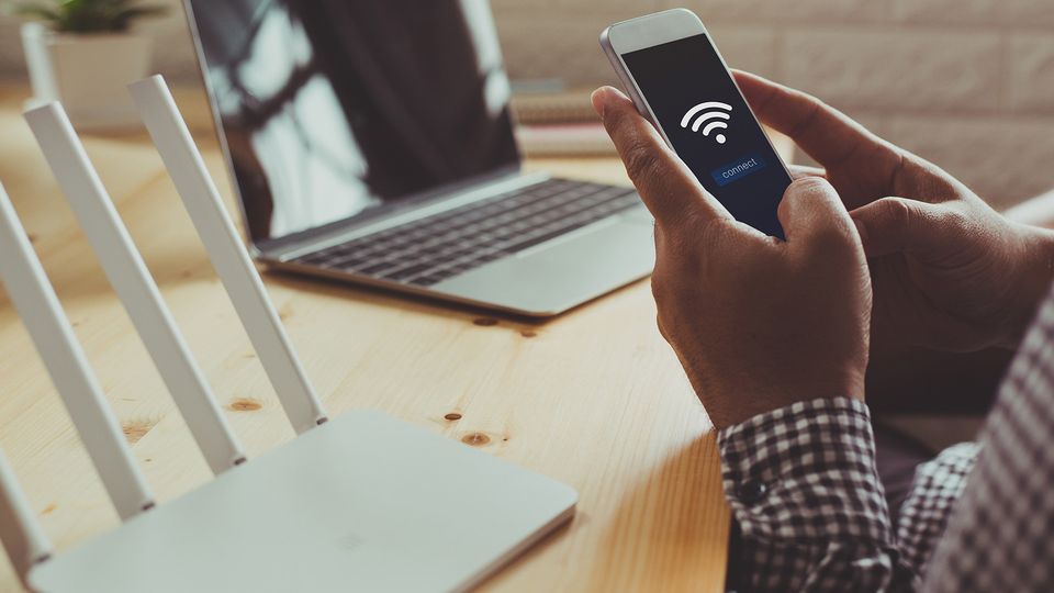 Bilde av en person som setter opp et wifi-nettverk hjemme med pc i bakgrunnen og mobilen i hånda.